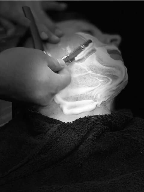 A client reclined and lathered, razor in hand, in black and white.