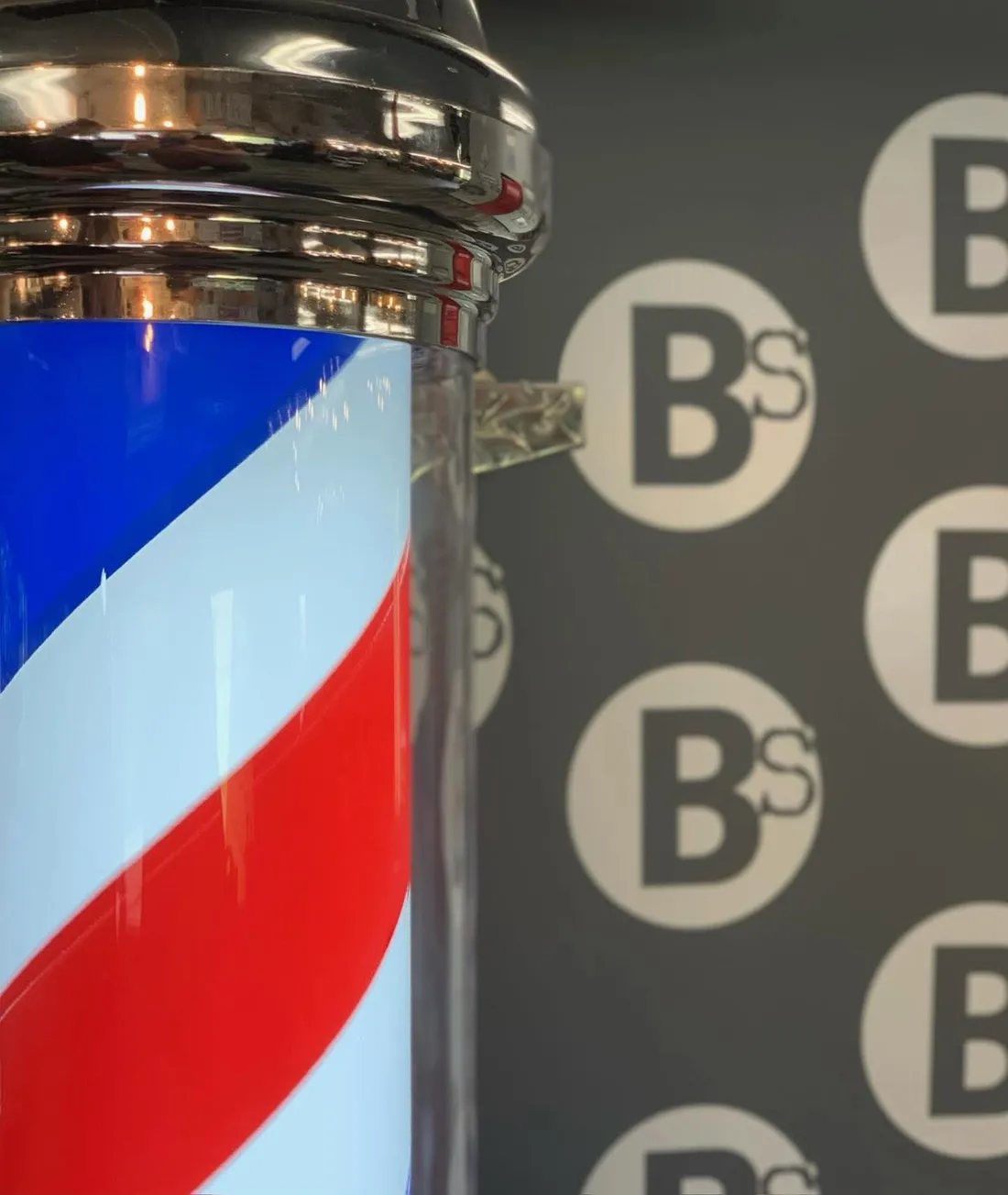A classic red, white, and blue barber pole beside the Barber Shack BS logo wall.
