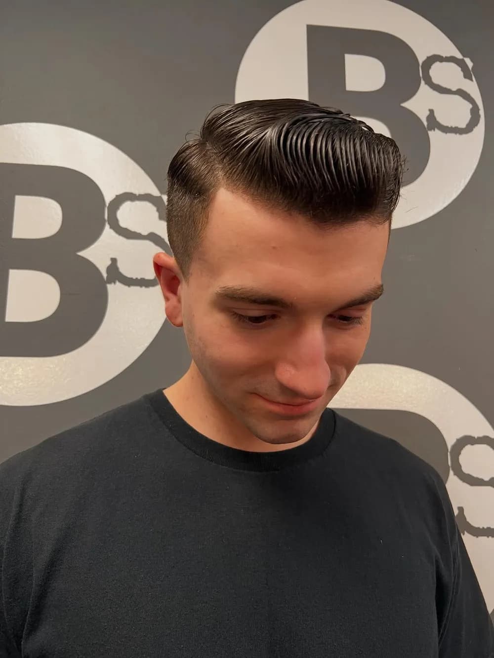 A fresh side-part pompadour cut and styled at Barber Shack, in front of the BS logo wall.